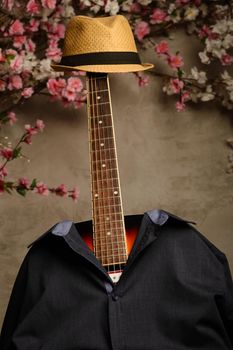 guitar is dressed in a man's blue shirt and a hat with a fretboard near a wall of flowers.