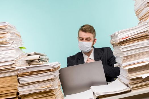A male accountant or company manager works in an office in a pandemic in view of the accumulated paper work. A protective medical mask is on the face. On the desktop are large stacks of documents. - image