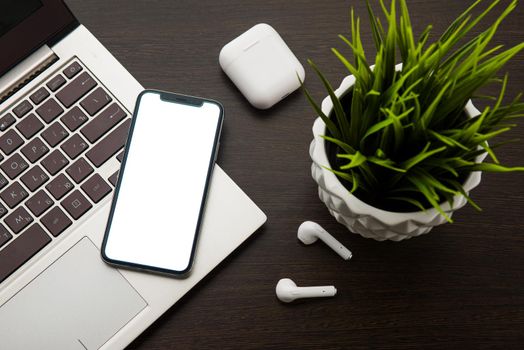 Top view on a smartphone laptop keyboard and wireless headphones on a brown table next to a houseplant - image