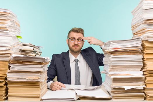 Shocked businessman sitting at the table with many papers in office, he is overloaded with work - image