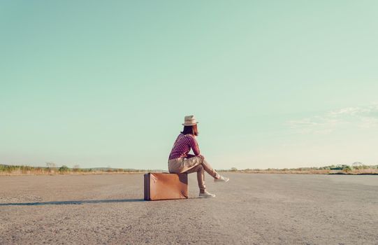 Traveler woman sits on retro suitcase and looks away on road, face is not visible