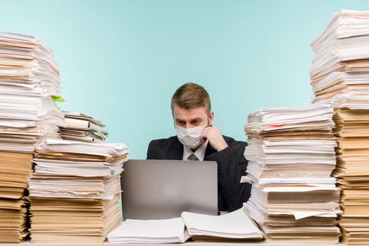 A male accountant or company manager works in an office in a pandemic in view of the accumulated paper work. A protective medical mask is on the face. On the desktop are large stacks of documents. - image