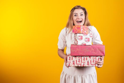 Portrait of a happy open-mouthed girl in dress holding gift boxes isolated over yellow background.