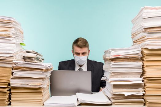 A male accountant or company manager works in an office in a pandemic in view of the accumulated paper work. A protective medical mask is on the face. On the desktop are large stacks of documents. - image