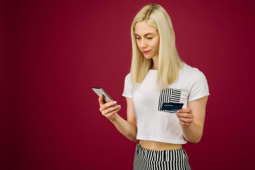 Young smiling woman buys online. Holds a smartphone and a credit card in hand on ruby background