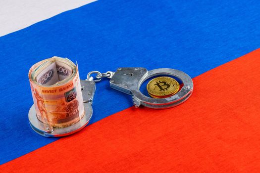 roll of russian paper rubles currency and bitcoin shiner chained with handcuffs on russian flag background - crypto ban law concept