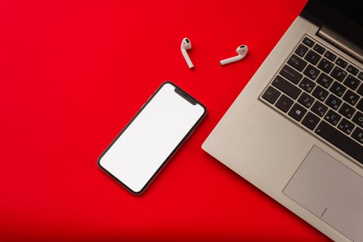 Tula, Russia - May 24,2019: Apple iPhone X and Airpods on red background with notebook. The screen of the smartphone is white. Mockup.