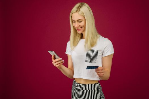 Young smiling woman buys online. Holds a smartphone and a credit card in hand on ruby background