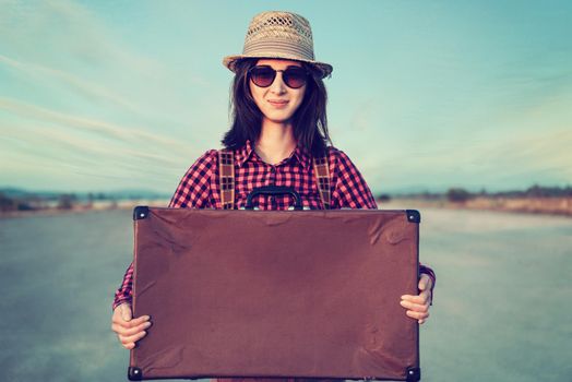 Smiling hipster traveler woman holds retro suitcase on road, space for text, theme of travel