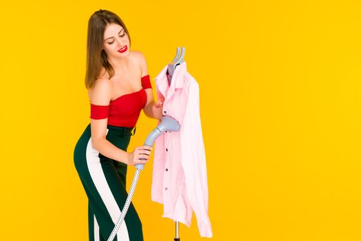 Young woman steaming pink shirt at dry-cleaner's on yellow background