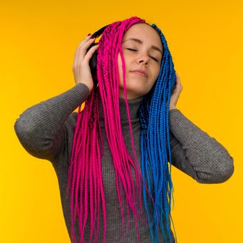 Happy cheerful woman with senegalese braids and freckles wearing wireless headphones listening to music from smartphone studio shot isolated on yellow background