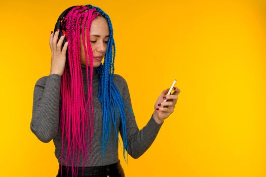 Happy cheerful woman with senegalese braids and freckles wearing wireless headphones listening to music from smartphone studio shot isolated on yellow background