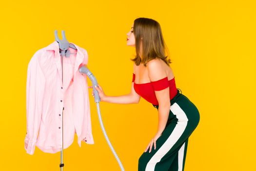 Young woman steaming pink shirt at dry-cleaner's on yellow background