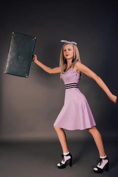 A young slender girl in a pink dress in high heels is hurrying with a suitcase in her hands on a journey on a dark background. Studio shot. Thin girl tourist.