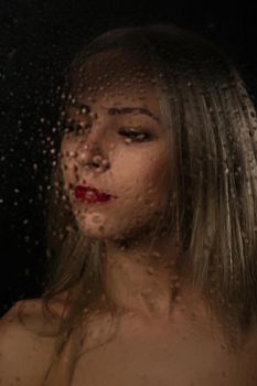 Smooth portrait of sexy model, posing behind transparent glass covered by water drops.