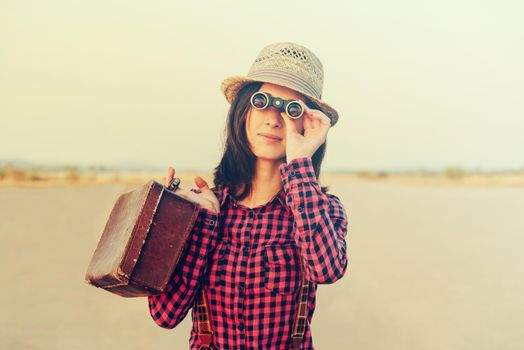 Tourist young woman with retro suitcase looks through binoculars outdoor. With vintage filter