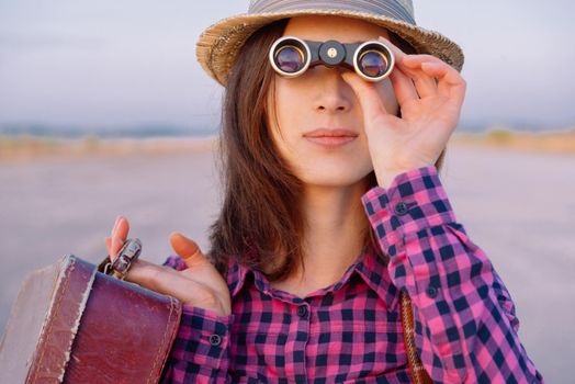 Young hipster woman with suitcase looks through binoculars