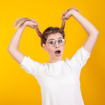 A young girl in glasses with her mouth open open in surprise holds her tails isolated on a yellow background