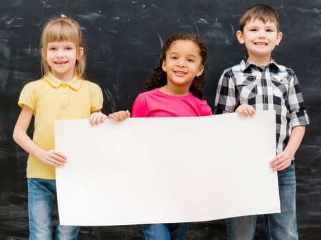 three cute children holding an empty paper sheet for ad with chalky blackboard on background