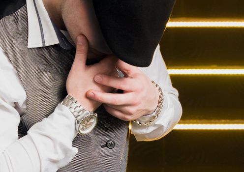 Fashionable hipster guy in a black flat cap beret and white shirt with a vest posing holding hands with a wristwatch and a schain over his face, close up.