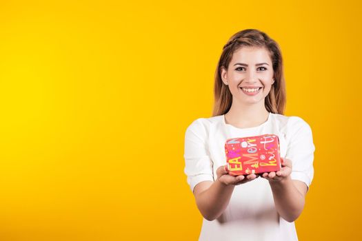 Portrait of a happy girl dressed in white dress holding little gift box and looking at camera isolated over yellow background