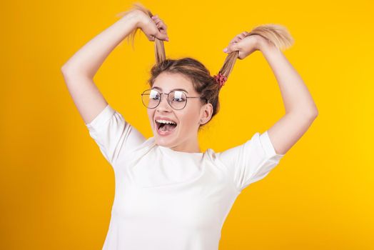 A young girl in glasses smiles and keeps herself behind tails isolated on a yellow background