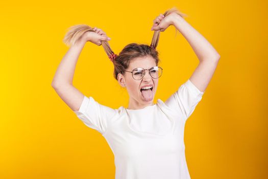 Young girl in glasses shows tongue and keeps herself by ponytails isolated on yellow background