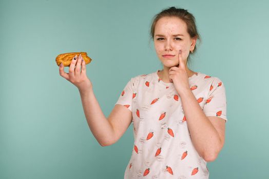 Portrait of pimply teen girl with cake points to acne on face on blue background