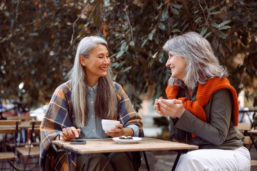 Positive mature Asian woman talks to best friend resting together at small wooden table in street cafe on nice autumn day