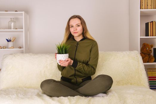Young teen girl with acne face sitting on sofa and holding green houseplant - image