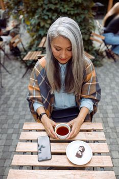 Tranquil long haired senior Asian woman holds cup of fresh tea sitting at table on outdoors cafe terrace on autumn day