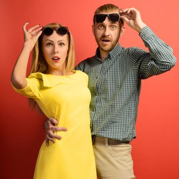 charismatic man in a green checkered shirt and a beautiful slender young woman in a yellow dress standing up with sunglasses and opening her mouth with surprise on a red background