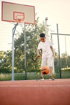 Young boy standing on basketball court near the park. Boy wearing white t-shirt. Boy playing with basketball ball.