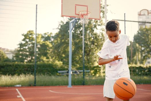 Young boy standing on basketball court near the park. Boy wearing white t-shirt. Boy playing with basketball ball.