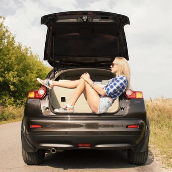 Young attractive woman sitting in the open trunk of a car. Summer road trip