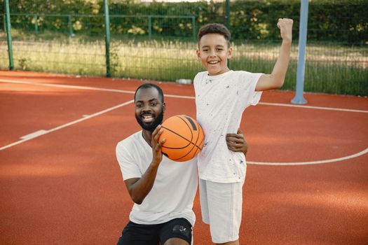 Young father and his son standing on basketball court near the park. Man and boy wearing white t-shirts. Boy posing for a photo pretending he is a winner.