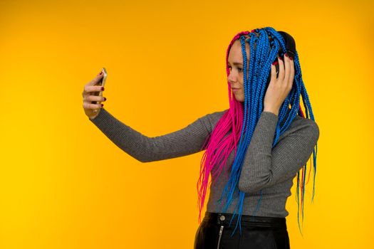 Happy cheerful woman with senegalese braids and freckles wearing wireless headphones listening to music from smartphone studio shot isolated on yellow background