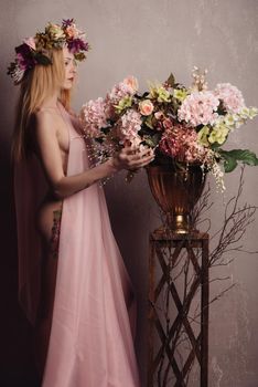 Beauty Summer model girl with colorful flowers wreath in a transparent pink fabric on the body stands next to a pot of flowers on a wooden box.