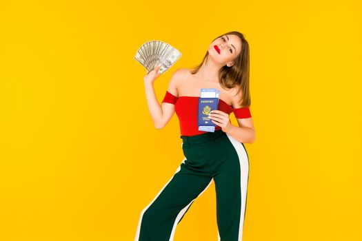 A young woman holds a money and passport with airline tickets. . Stylish girl on in a red blouse and green pants on a yellow background. - image