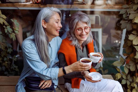 Smiling mature Asian lady with friend clink cups of coffee sitting on bench near large cafe window outdoors. Long-time friendship