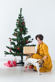 Happy woman with dog opening Christmas gifts. Christmas tree with presents under it. Decorated living room.