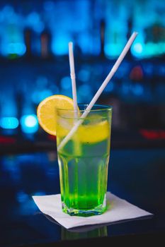 Cooling alcoholic cocktail with green syrup and lemon on a bar counter against a blue nightclub background.