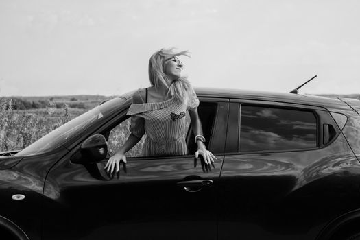 The thin girl got out of the car window and smiles at the sun. Black and white