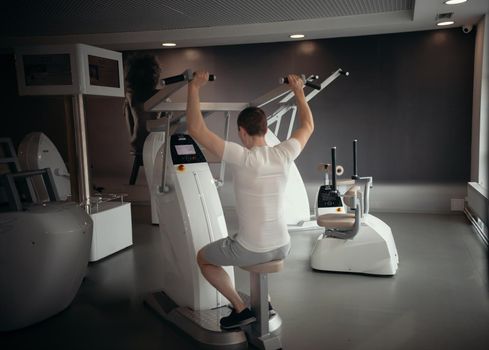 Modern gym - a young man in white t-shirt sitting on the strength training machine and pumping his hands muscles. Mid shot