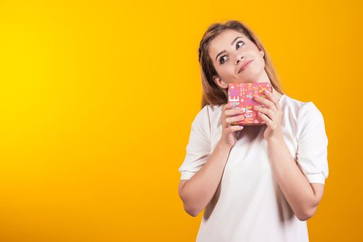 Portrait of a happy girl dressed in white dress holding little gift box and looking away isolated over yellow background