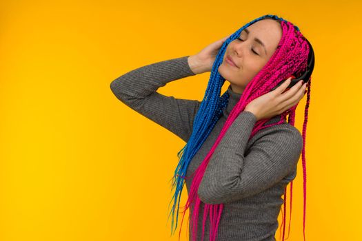 Happy cheerful woman with senegalese braids and freckles wearing wireless headphones listening to music from smartphone studio shot isolated on yellow background