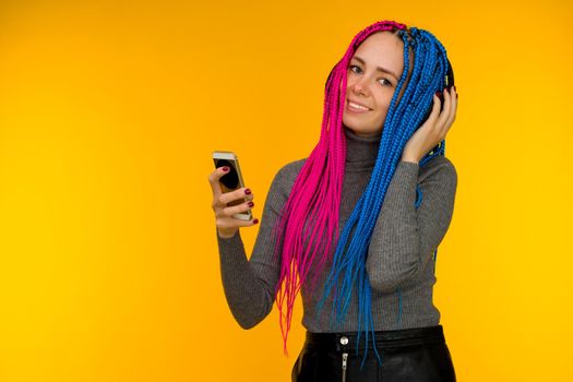 Happy cheerful woman with senegalese braids and freckles wearing wireless headphones listening to music from smartphone studio shot isolated on yellow background