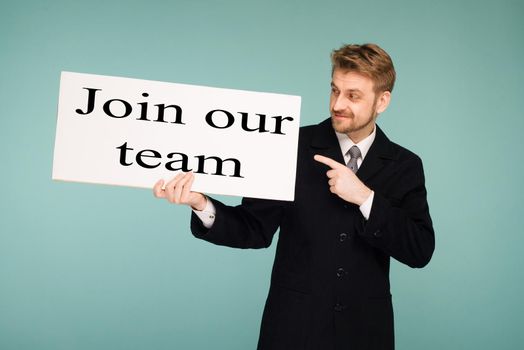 Happy smiling young business man points finger signboard with sign Join Our Team, on blue background