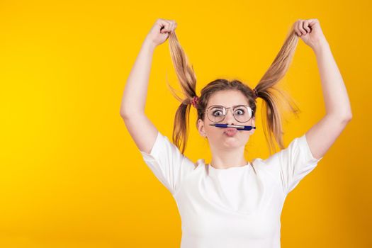 Young girl with glasses holds herself by the tails holding the pen lips isolated on a yellow background