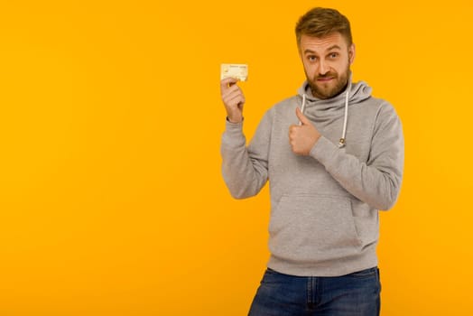 Attractive man in a gray sweatshirt raised a finger up holds a credit card in his hand on a yellow background - image